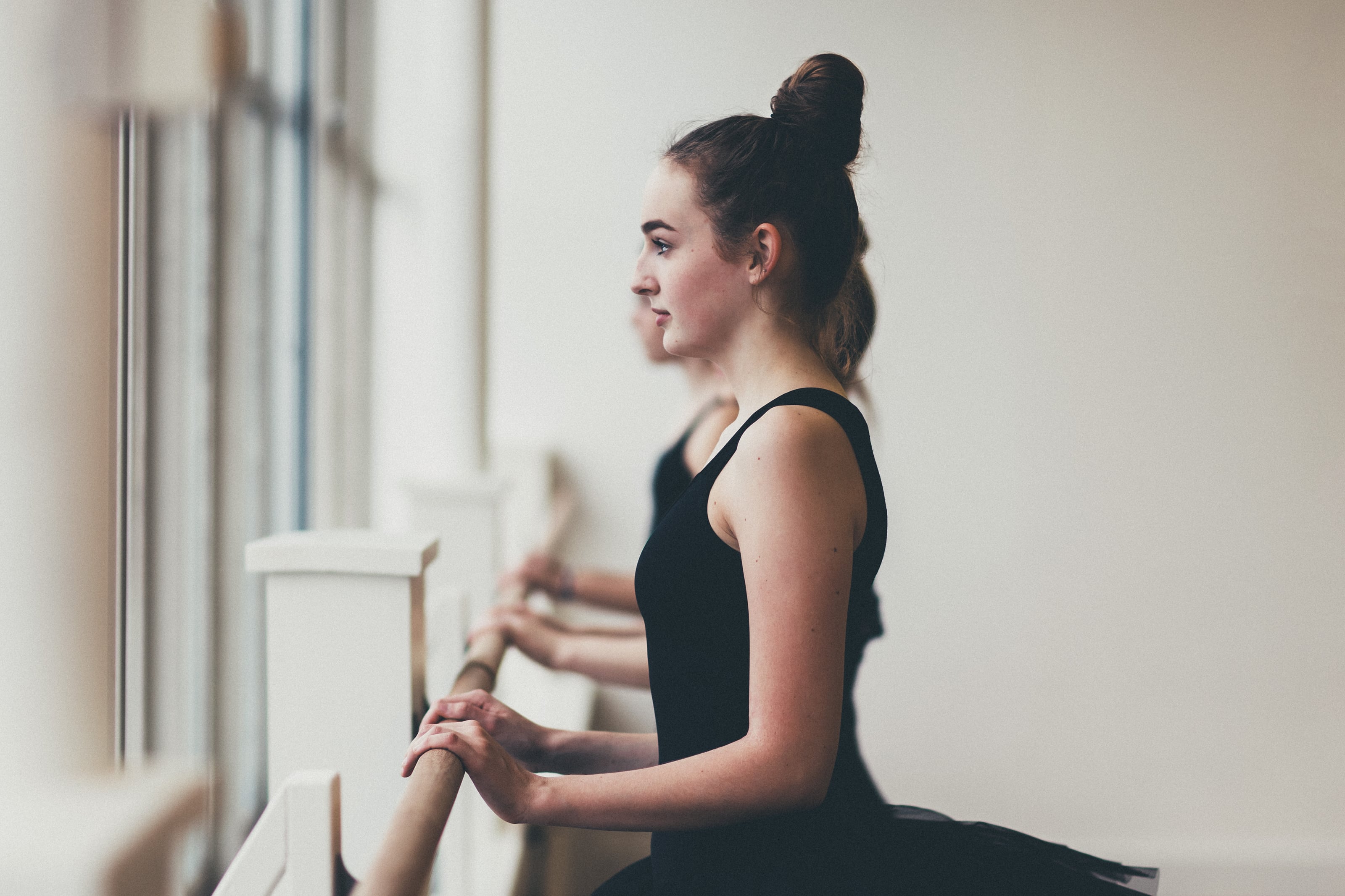 Dancer in black tutu stands at a barre beside tall windows.