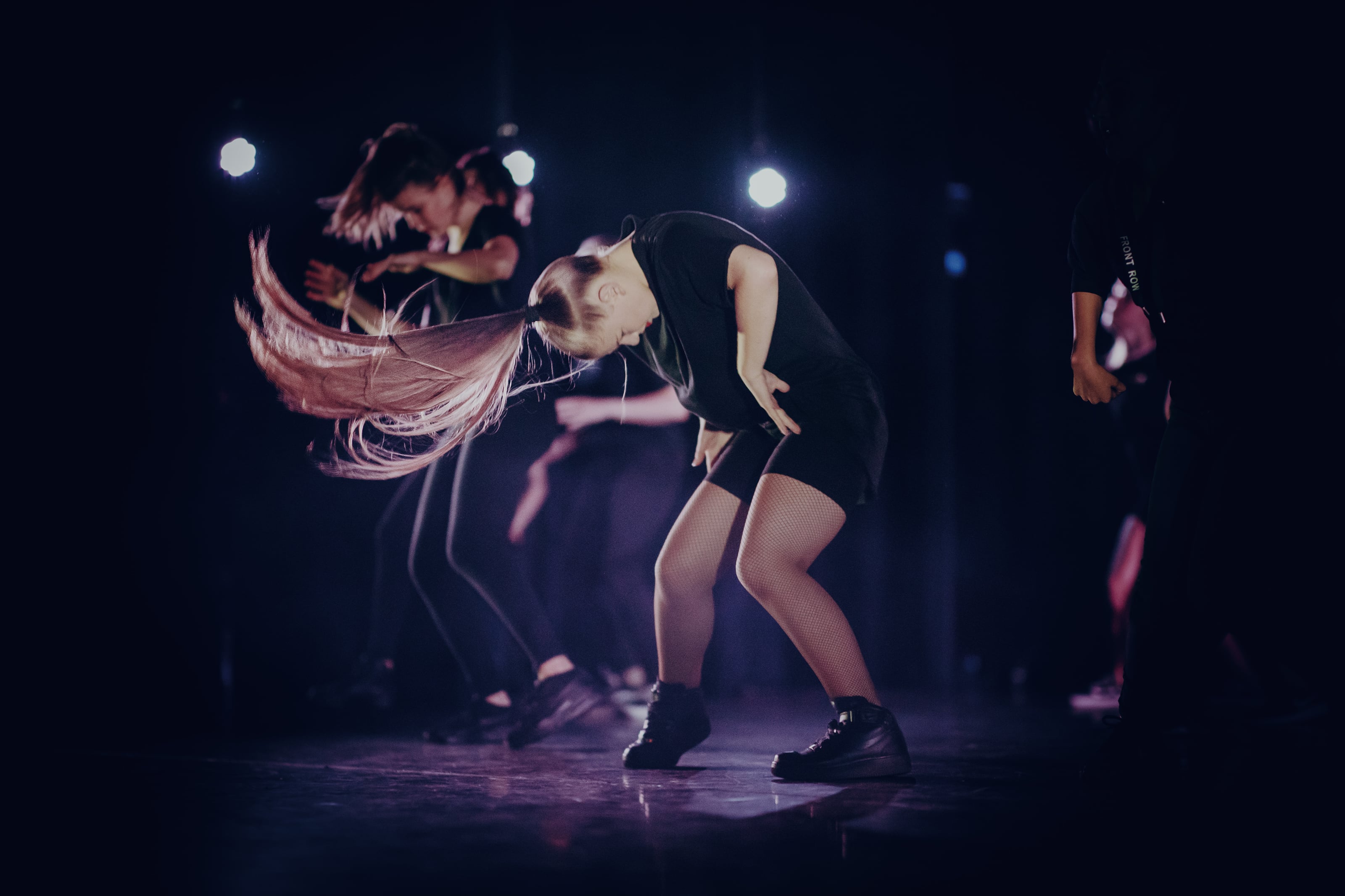Dancers perform a hair-whip move on a dark stage with spotlights.
