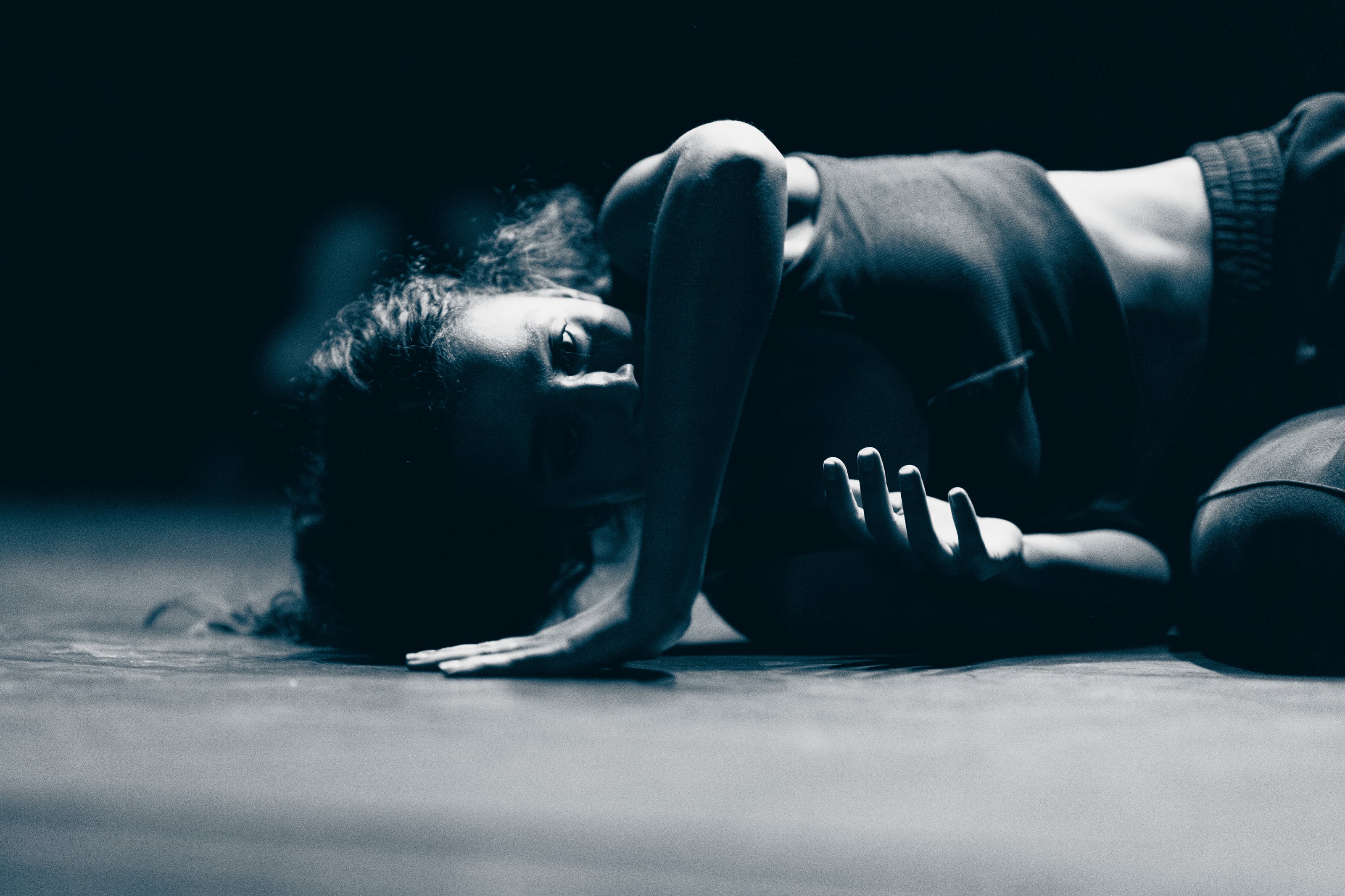 Person lies curled on a dark floor under dramatic side lighting.
