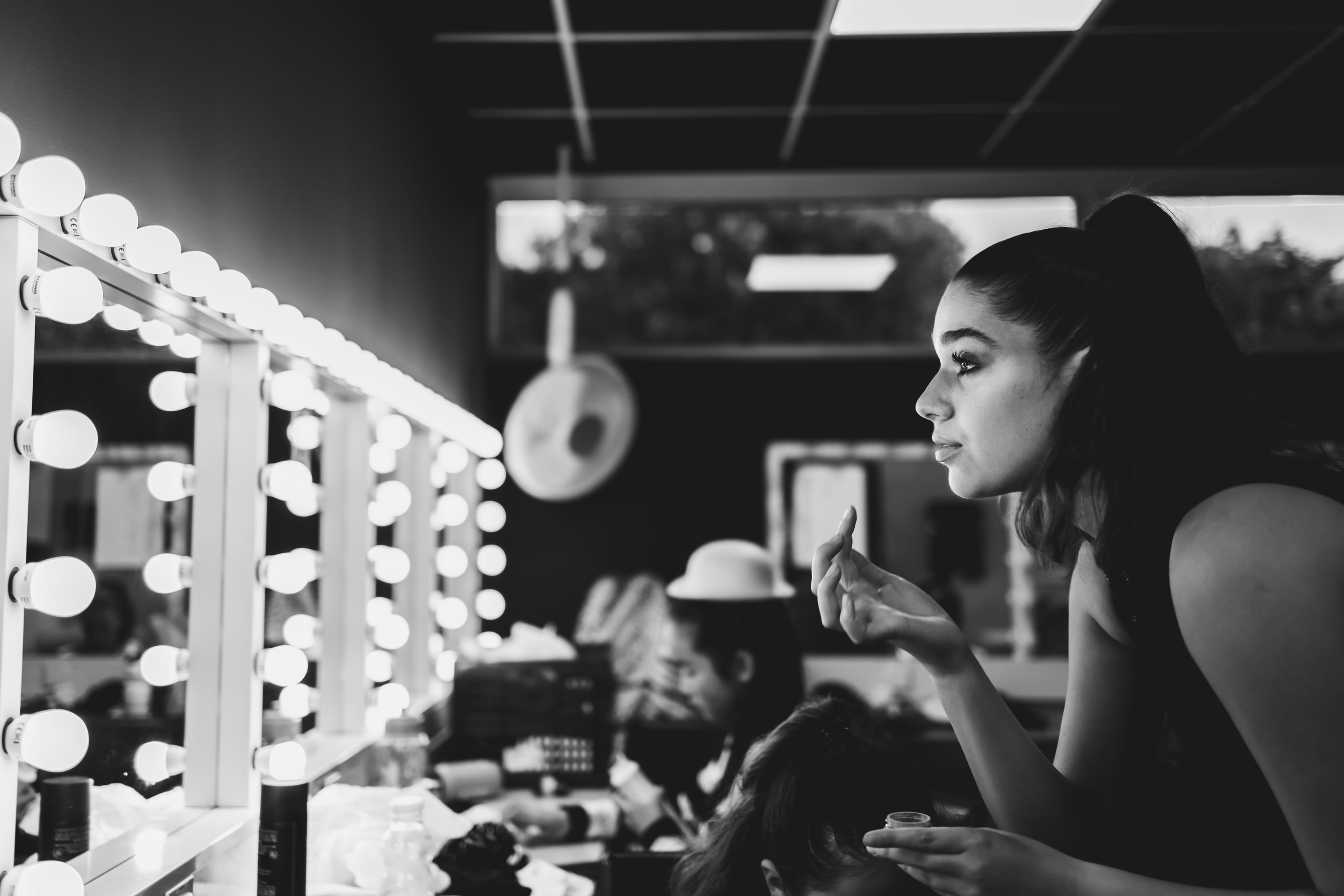 Woman applies makeup beside illuminated mirrors in a dressing room.